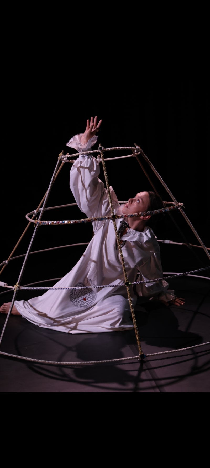 Image of a woman in an embroidered 16th century smock, reclining on the ground inisde a jeweled fathingale-cage, reaching her right hand upwards.