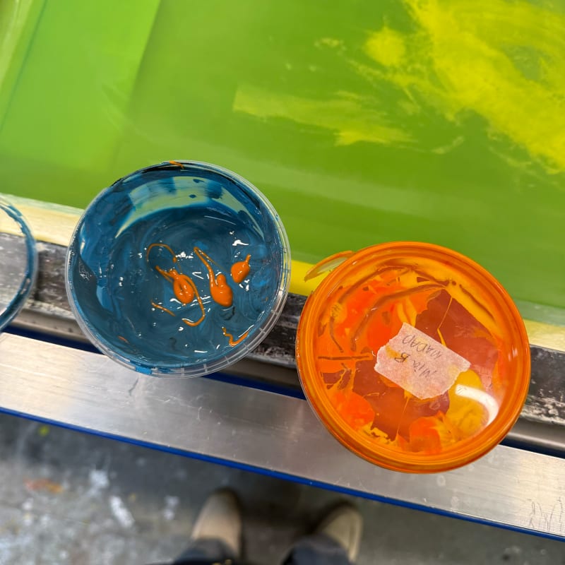 Two screen-printing ink pots, one blue and another orange, next to a silk-screen.