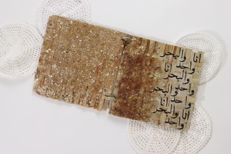 Woven palm leaf structures with embroidery of Arabic calligraphy 'the ocean and i are one' in a book format.