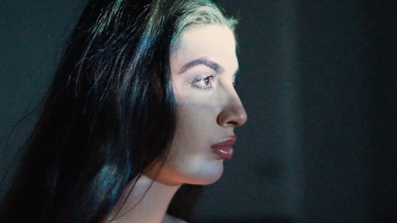 A close-up of my contributor, Aruzu, looking directly at a projected image of her younger self dancing, the projector’s glow illuminating her face.