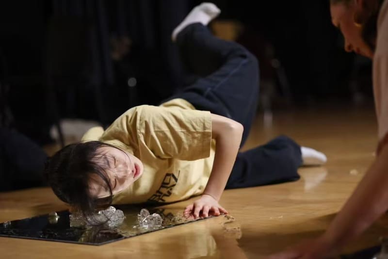 Dancer posed on a stage in a sideways manoeuvre . Blocks of ice are melting on the stage and the dancer is posed with their face close to ice blocks on the floor.