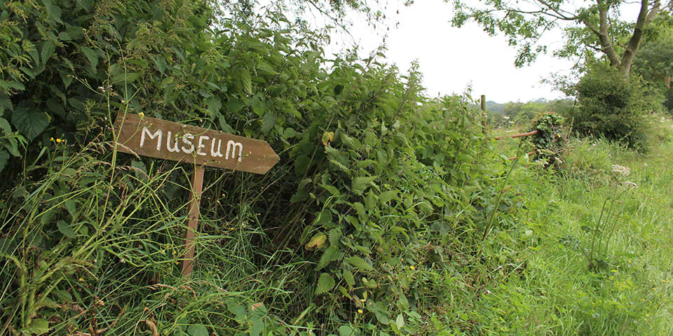 Photograph of a wooden arrow sign that reads 'Museum' in the middle of woods.