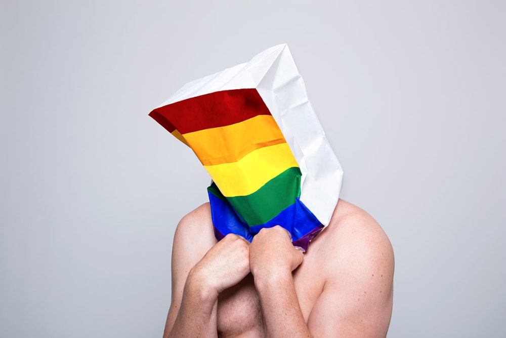 Photograph of a man wearing a paper bag with the LGBT rainbow flag on it by Christa Holka