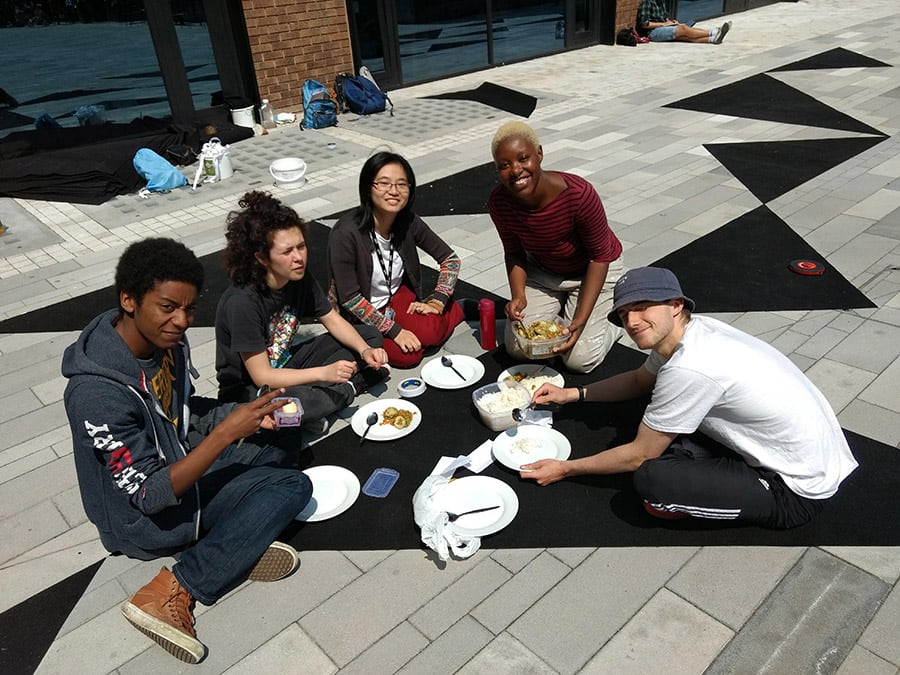 A group of students eating a picnic on the black triangle patterned floor outside. 