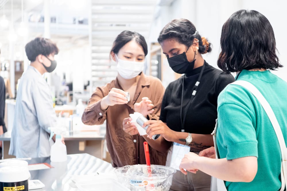 Nathini, centre, has two people either side of her. They are all wearing masks and appear to be in a kitchen. There is a glass bowl on the table, covered with cling film and they are holding a silver tube.