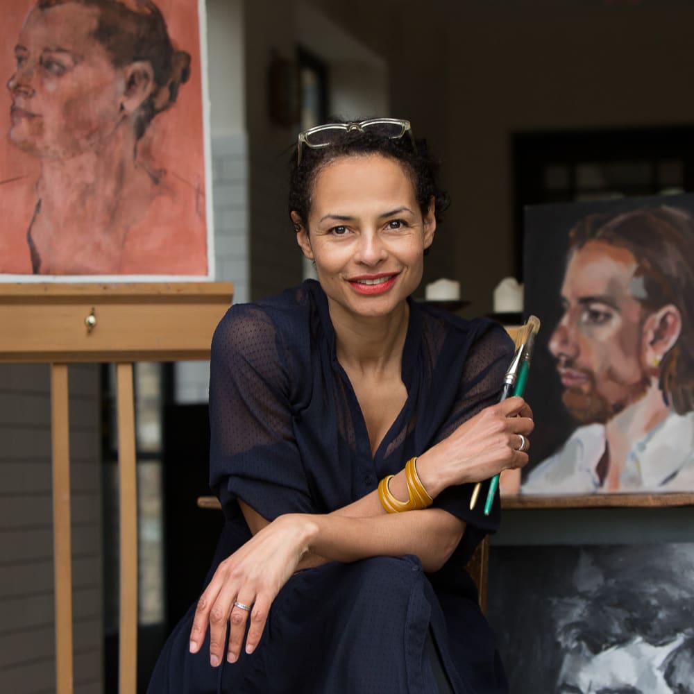 Photo of Jacqueline smiling to the camera. She's holding paint brushes and is surrounded by her work. 