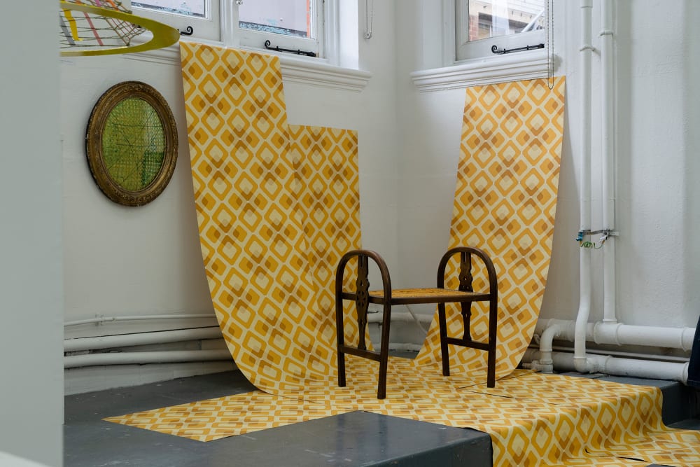 A patterned design is hung down a wall and across the floor, on the design stands a dark brown wooden bench and a round brassed frame weave is displayed on the wall. 