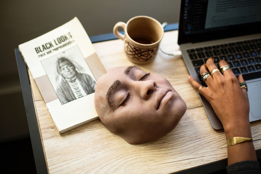 A body cast of a face lays on a table on which we see a hand typing on a laptop.
