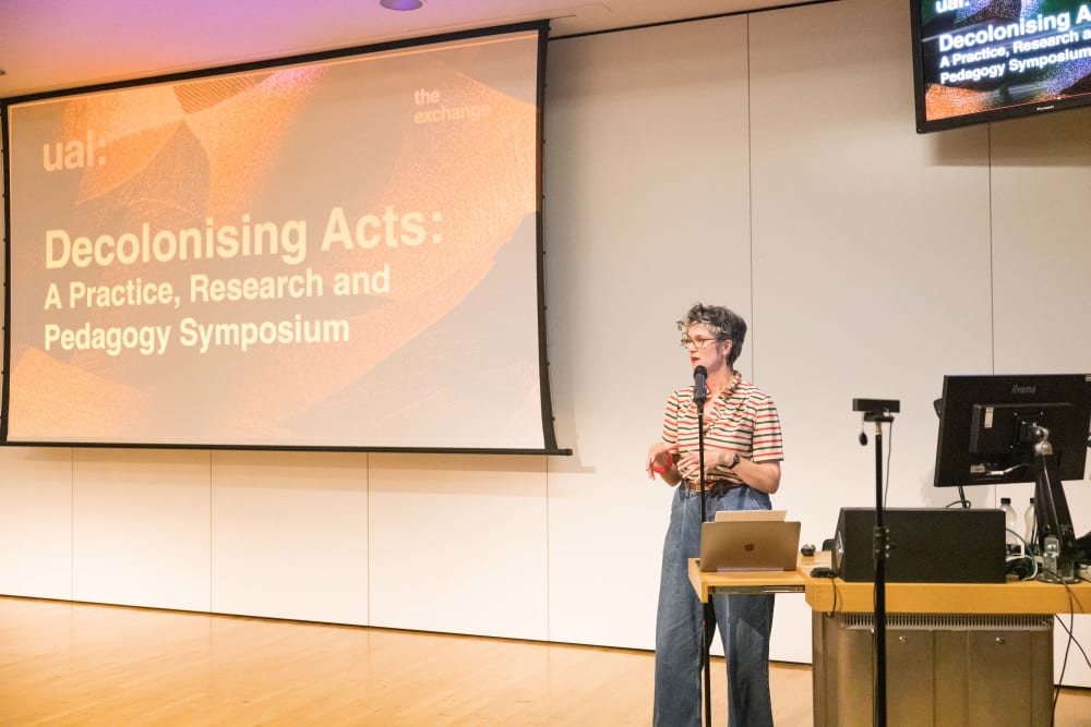 Standing woman speaks into mic at front of event hall. 