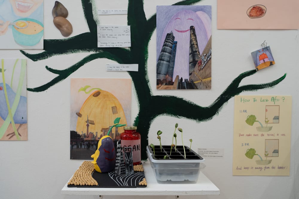 An abstract artwork is hanging against a white wall. On the left the material is ruffled with blue twine weaved through. On the right a whole piece of material with dark greens, blues and browns interweaved with pebbles scattered on top.