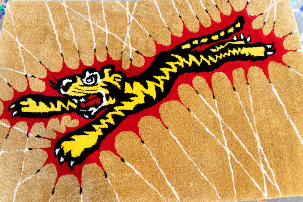 Detail of work by Graphic Design student Charlie Calvey. This is a carpet showing a cartoon tiger surrounded by a red jagged outline on a tan background with white stripes coming from the points of the red outline.