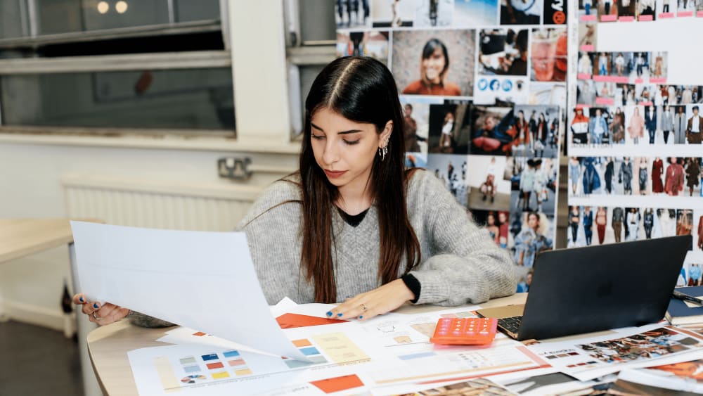 A woman with long dark hair sits at a desk, reviewing a large, printed sheet. Her workspace is filled with papers, a laptop, a red calculator, and colorful fashion-related materials. Behind her is a wall covered with fashion inspiration images and mood boards.