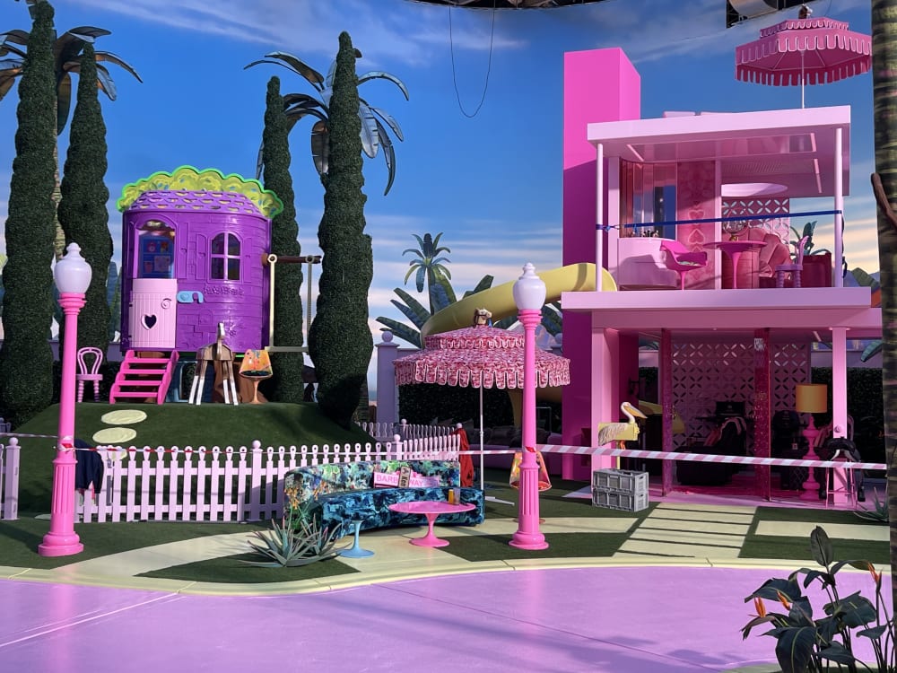 Image shows a purple trailer house on the left-hand side, surrounded by green trees. On the right-hand side, there is a pink house. In the background you can see a blue skyline and in the foreground is pink paving.