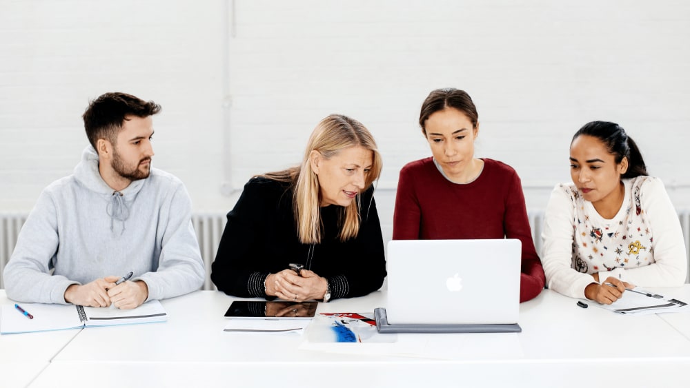 Four people, three women and one man, sit at a white table in a bright room, focused on a laptop screen. They appear to be collaborating or in a meeting. The table has notebooks, papers, a tablet, and pens.