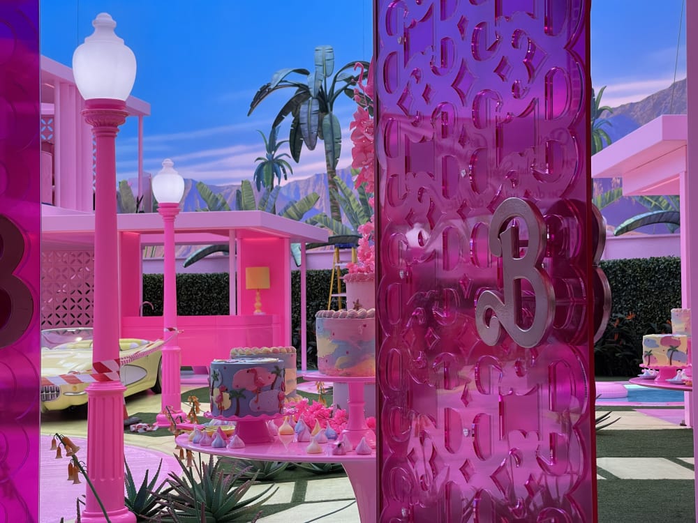 Image shows a pink door with the letter ‘B’ acting as a door handle. You can see a pink lamppost through the door, along with a table with cakes on top. In the background are palm trees and a blue sky line.