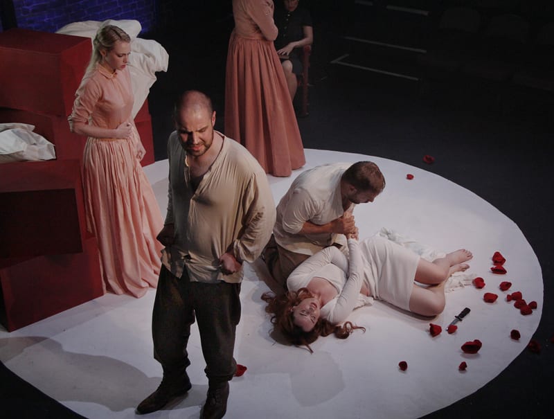 A stage set with a woman lying on the floor, a man sitting over her holding her hand and three other figures surrounding her. 