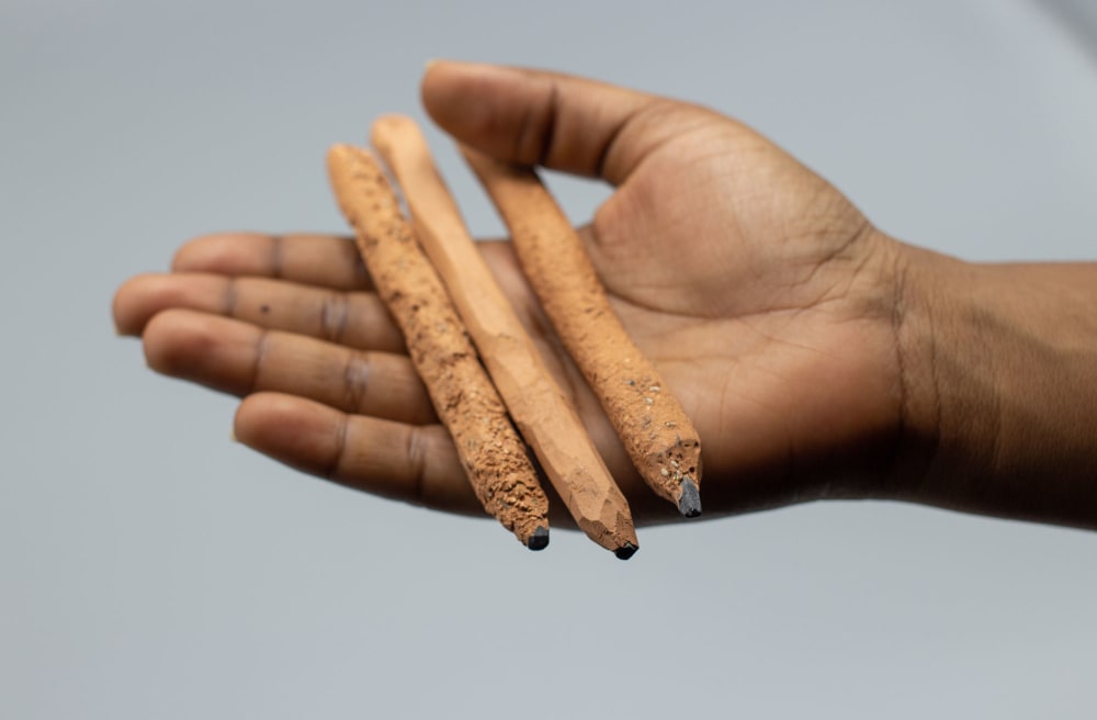 A hand holds three pencils which appear to be crudely carved from wooden sticks.