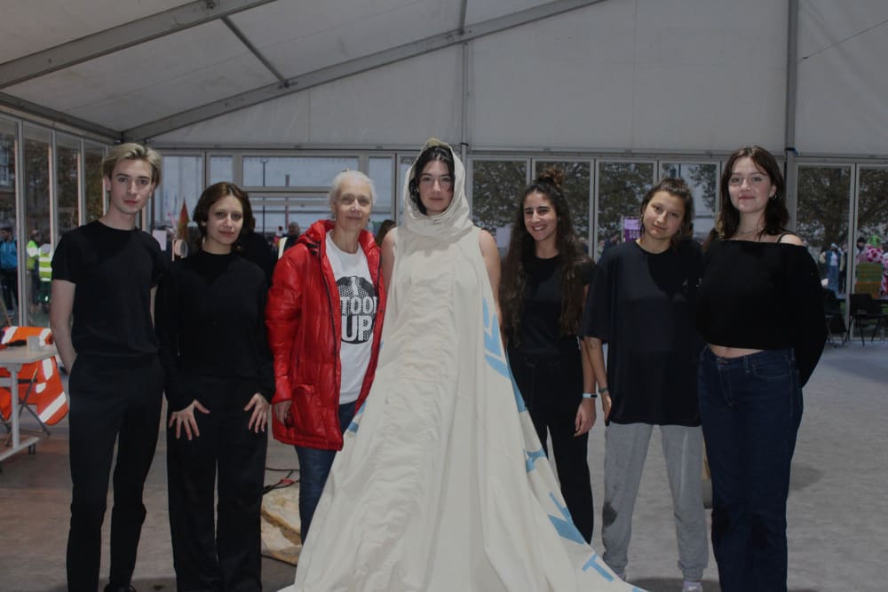 A group of 7 pople standing in a tent-like room looking at the camera. At the centre of the group is a woman in a white canvas dress and hood, next to a woman in a red jacket. The other people all wear black.