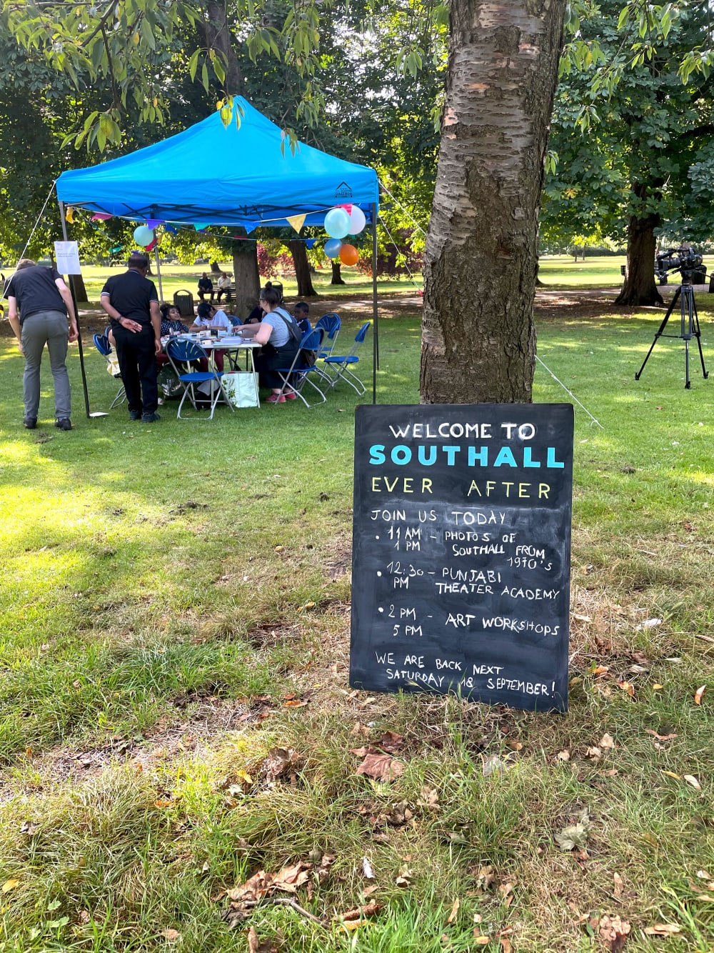 A blackboard sign saying 'Welcome to Southall Ever After' leans against a tree in a park with a gazebo visible in the distance behind it.