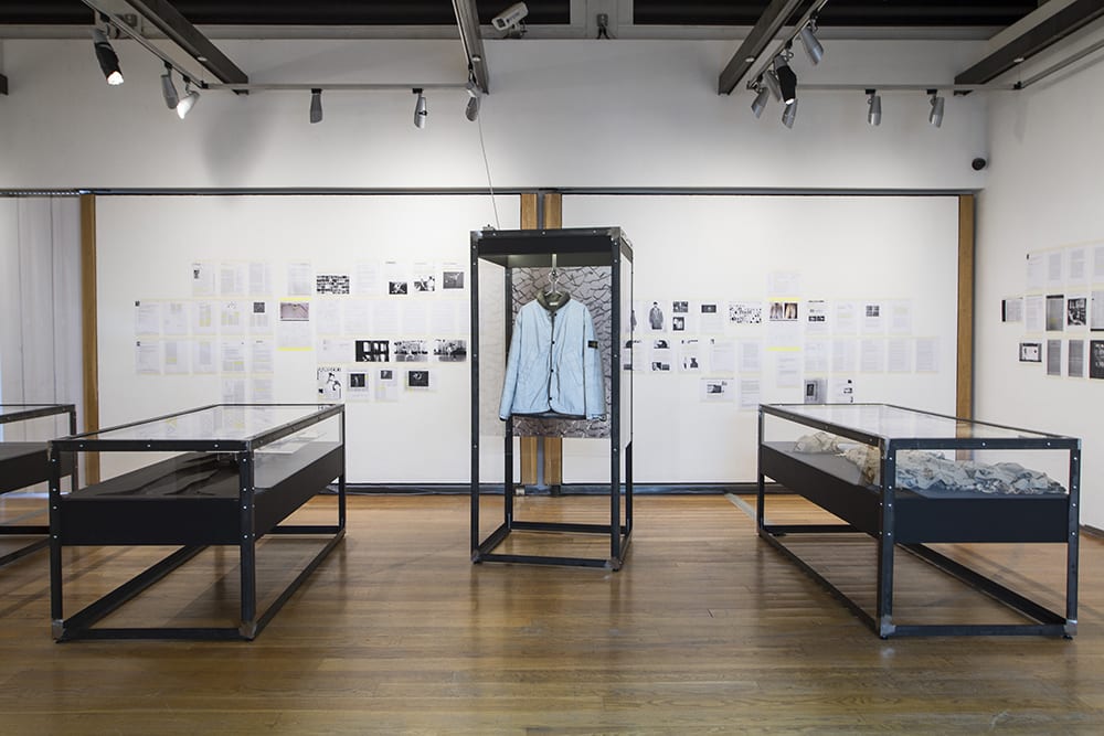 Exhibition space containing upright glass case containing jacket with horizontal glass cases either side