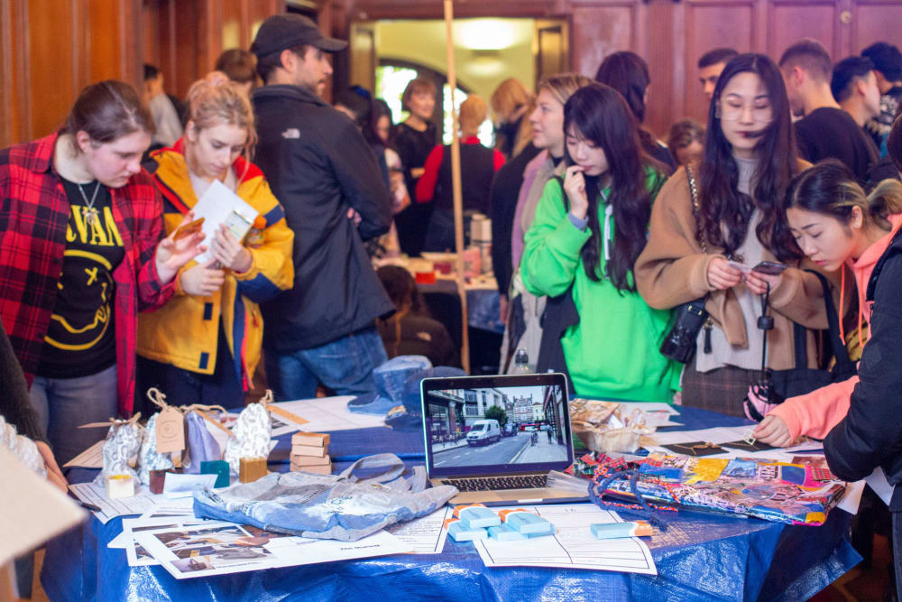 A busy room of people surrounds a table which displays work, papers and a laptop computer.