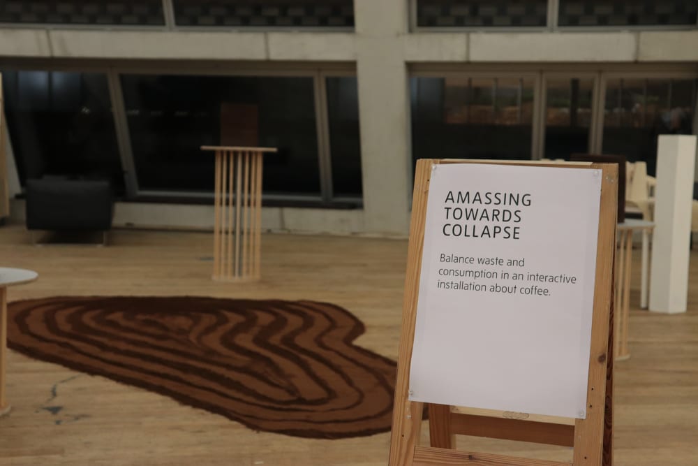 Inside the exhibition space at the Tate Modern, there is a wooden A board with a sign that says ' Amassing Towards Collapse' behind the board is 2 podiums of coffee and there is a coffee rug on the floor. 