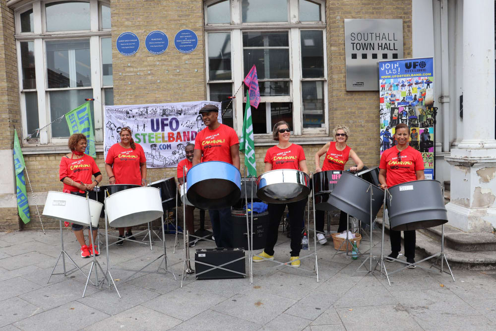 7 people in red t-shirts playing steel pans in front of a yellow brick building.