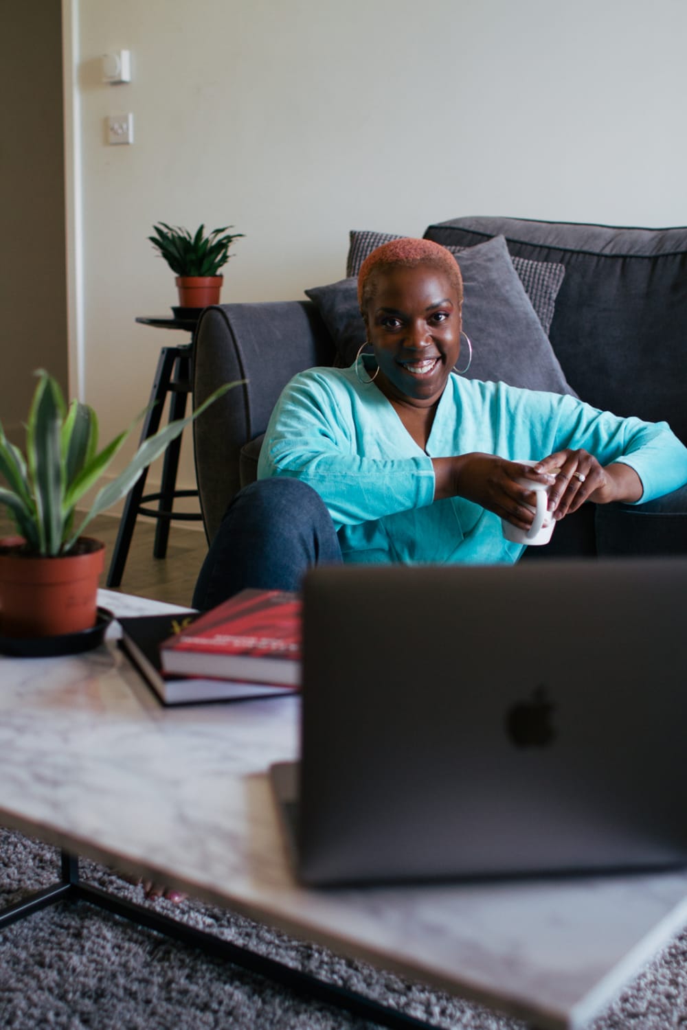 Danielle is sat on the floor in front of a sofa, and behind a coffee table. There's a laptop on the table next to her. Danielle is wearing a blue shirt and smiling into the camera.