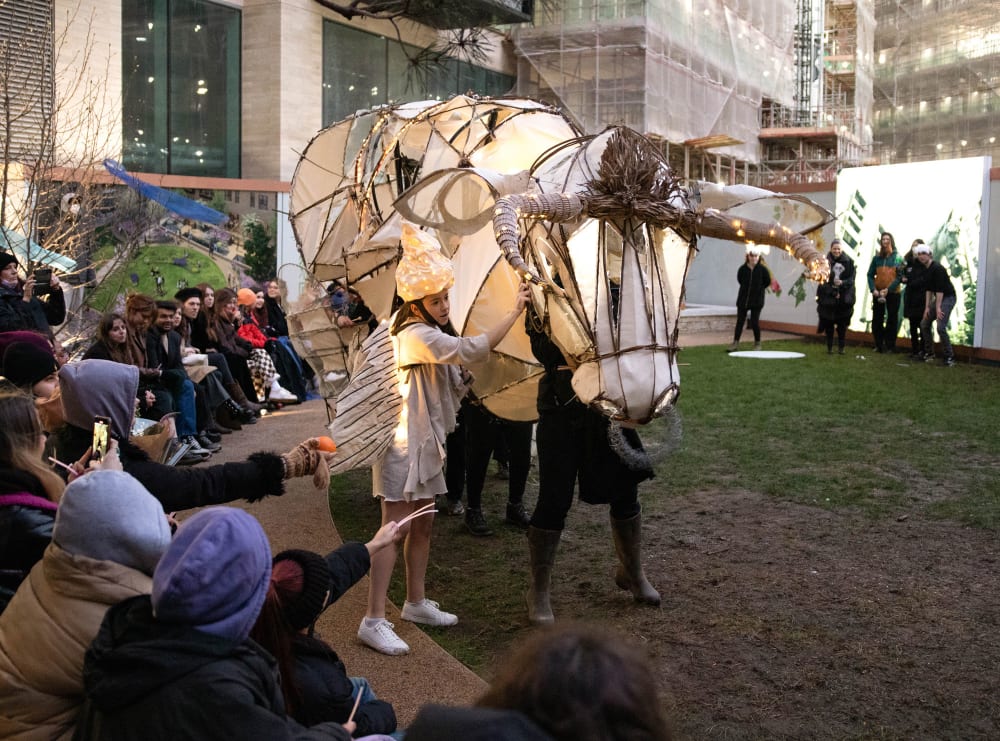 A large white paper puppet of a bull, operated with a person inside it, performs next to a woman dressed in silver with a light-up shell headdress.