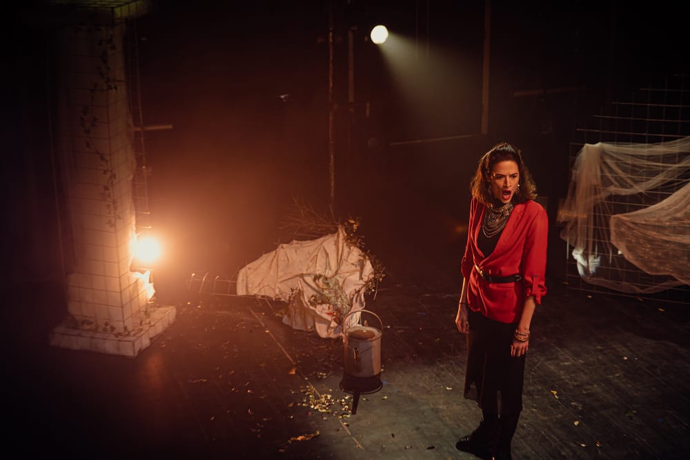 A woman in a red coat looks to be shouting, she faces forward. The stage is set,  there is a watering can on the floor and white sheets draped over metal wire messed with leaves scattered. 