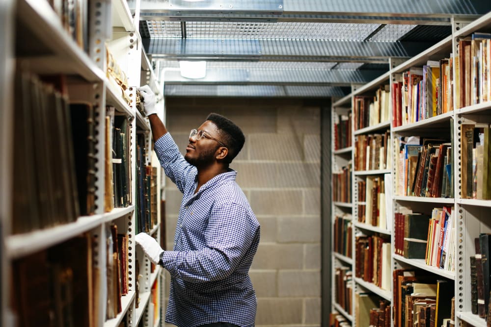  A man in a checkered shirt and white gloves reaches for a book on a tall shelf in a library archive.