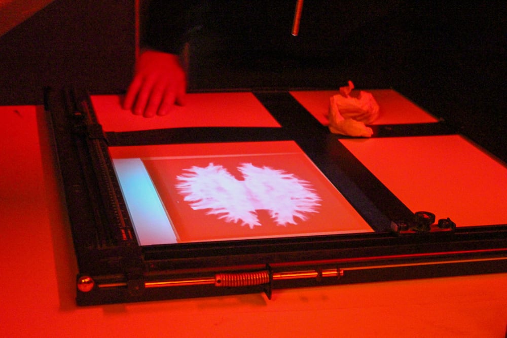 The photograph taken in a dark room has a red tint, there is a hand placed on a negative of a brain scan which is being lit up on a light box. 