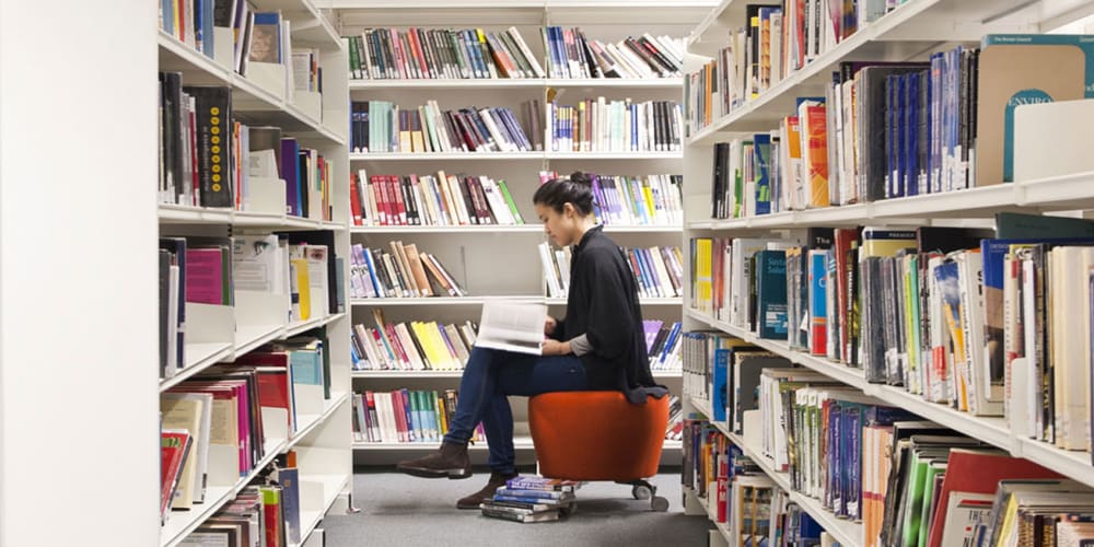 Photo of a girl sat in between two book shelves reading