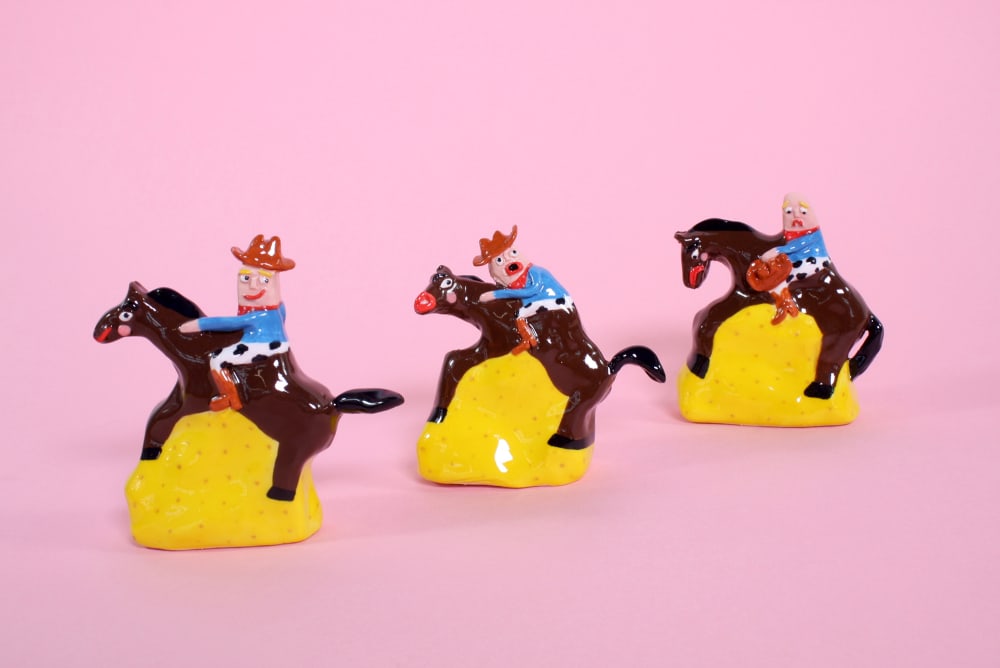 Three ceramic figurines of cowboys riding horses. The brown horses stand on yellow and are photographed against a pink background. Each of the cowboys has a different facial expression: (from l-r) Happy, surprised and sad.