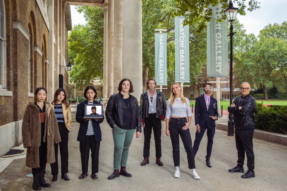 8 people standing outside the Saatchi gallery - they are the curators of the exhibition. One person is holding a laptop open with a portrait of a woman on the screen - she is remotely curating. 