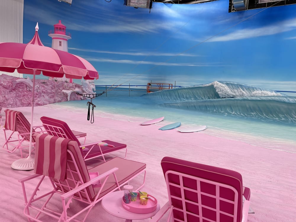 Image shows 4 pink deckchairs and a pink umbrella in the foreground sitting on top of a pink sand beach with blue water and skyline in the background. To the left of the image, a pink and white lighthouse can be seen sitting on top of pink rocks. 3 surfboards can be seen at the water’s edge.