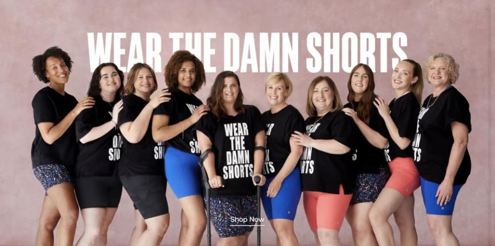 A group of women standing together in a long line wearing black t-shorts and shorts, standing in front of a pink wall with the words 