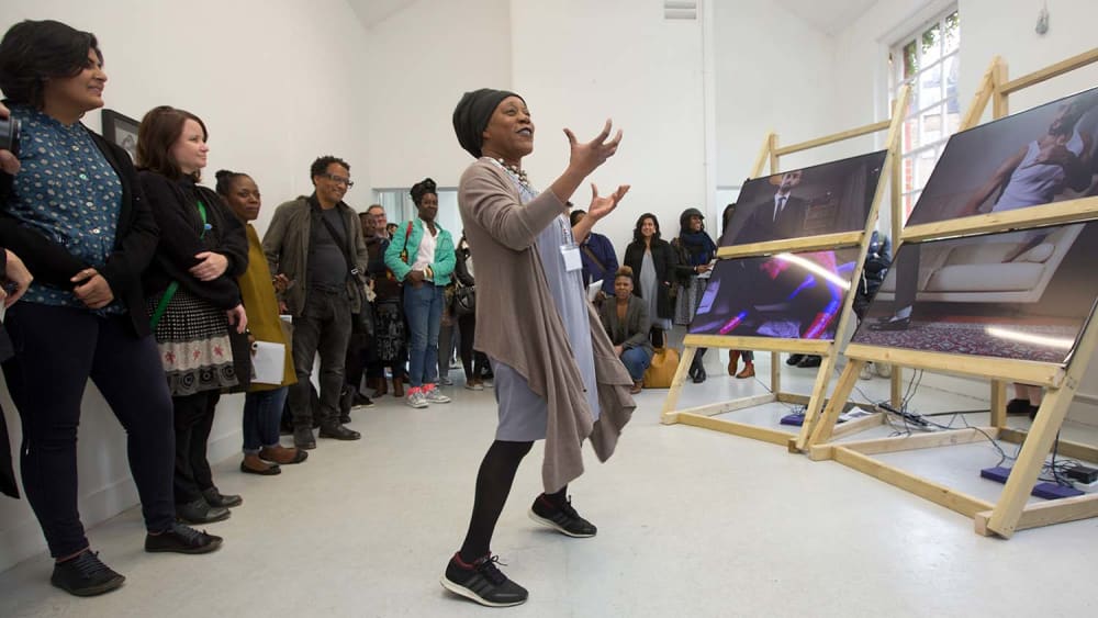 Sonia Boyce standing speaking with audience to left and video installation to the right.