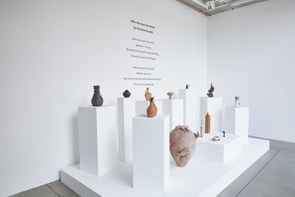  A side view of an exhibition, 8 white podiums each with a ceramic piece positioned on top surround a smaller but wider podium with 4 ceramic pieces positioned together. Next to the smaller podium a large ceramic vase is positioned. They are all sat on a white stage in front of a white wall, where the words of a poem are written in black lettering. The poem says: ‘Who Has Seen the Wind ? By Christina Rossetti. Who has seen the wind? Neither I nor you: But when the leaves hand trembling The wind is passing through. Who has seen the wind? Neither you or I: But when the trees bow down their heads, The wind is passing by.’