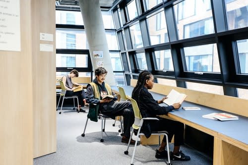 Students sitting at tables looking at books in front of large windows