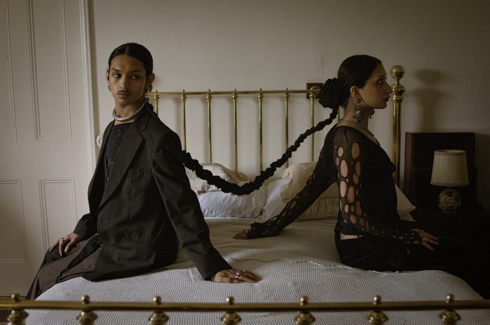 Two people with interlinked braids of hair sitting on a bed.