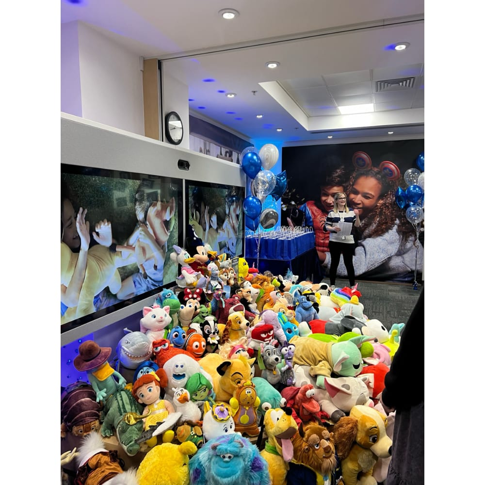 TV screens playing adverts behind a sea of soft toys of Disney film characters, at a Disney event. 