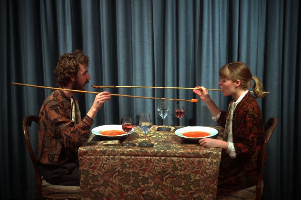 an image of 2 people sitting at a dinner table feeding each other soup with long bamboo spoons
