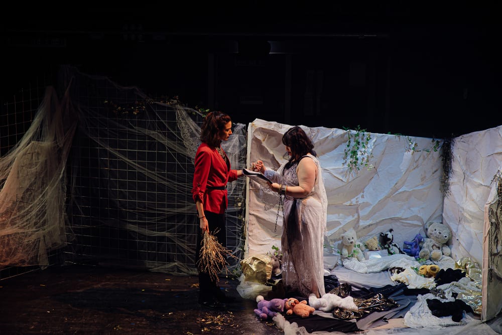 A stage set, 2 people stand facing each other holding hands. 1 is dressed in red, the other white with her wrists in chains. The woman in white stands among bed sheets and teddy bears with leaves scattered. 