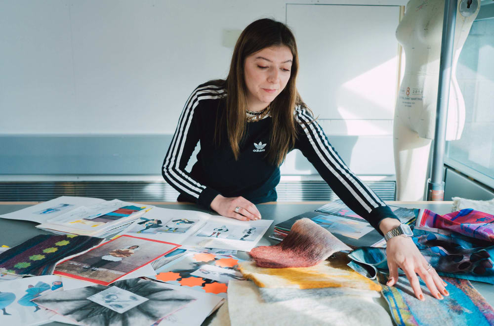 A woman in a black and white tracksuit leans over a table covered in sketches and fabric sampless.
