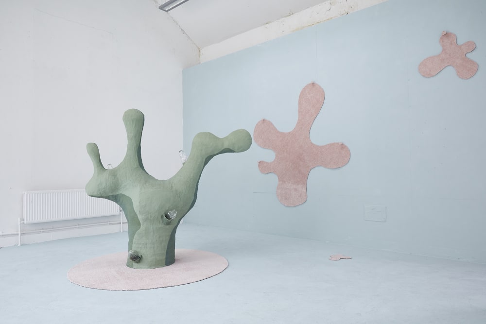 A green cactus looking sculpture stands on a large textured pink circle. The wall behind the sculpture and floor around the sculpture is painted in a light blue. On the wall there are two textured pink abstract shapes on the wall, they look as though they are splats of a paint, one is large, and one is small. 