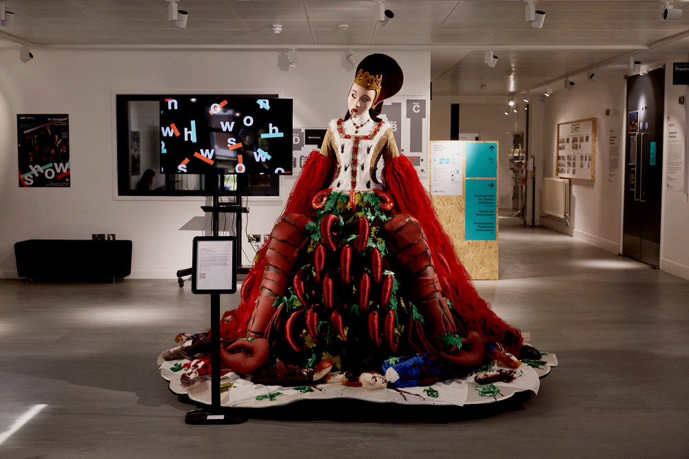 Image shows a mannequin with a crown on its head wearing a red lobster dress which has a ‘royal’ feel to it. The mannequin sits at the entrance to the College and you can see a TV screen in the background reading ‘Show’.