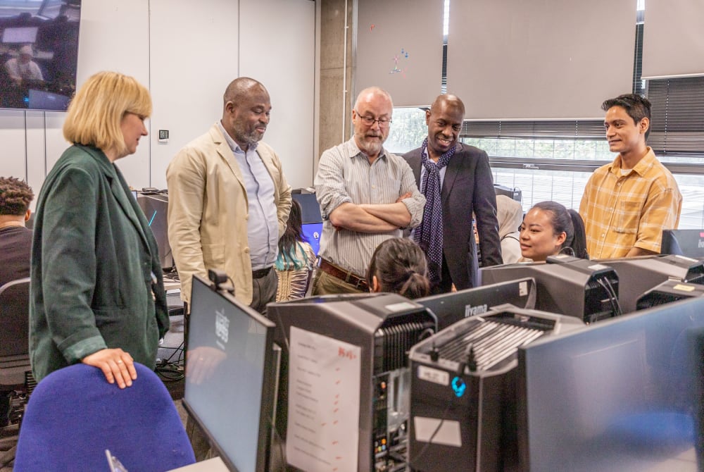 Students and staff standing around computers, engaged in discussion and collaboration.