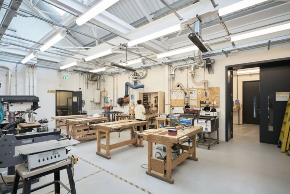 Interior of a workshop with wooden tool benches in the centre and machinery around the edges. The lighting is bright from ceiling lights and windows. There is a person standing at a cabinet looking at tools in the centre.