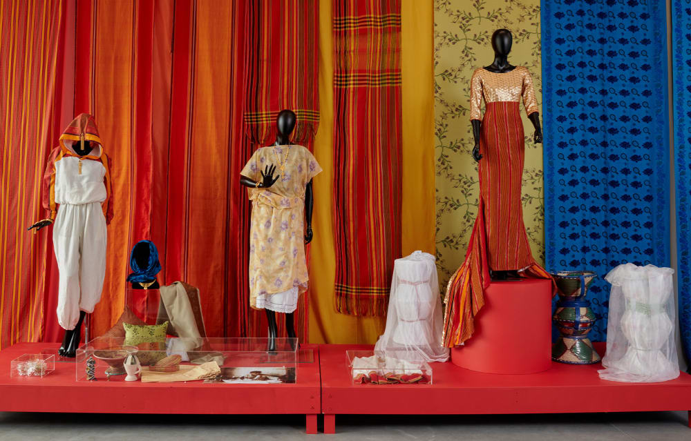 Three mannequins display colorful dresses against vibrant, patterned fabric backdrops, with accessories and textiles arranged on red platforms in front of them.